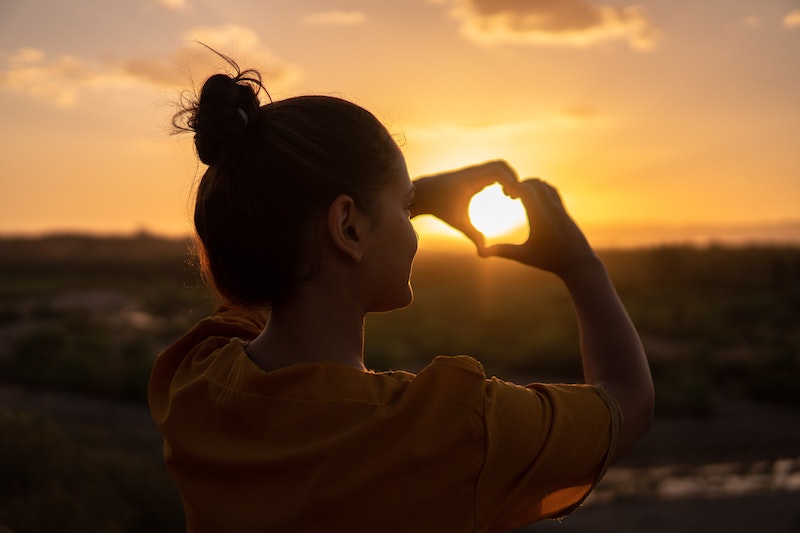 The silhouette of a woman is standing outside in front of a bright orange sunset. The woman has her hands lifted and has created the shape of a heart with her fingers, placed over the sun. The sunset shines through her hands. Contact trauma informed, EMDR and Somatic Experiencing trained therapists today. Mental health support available online across Ontario.