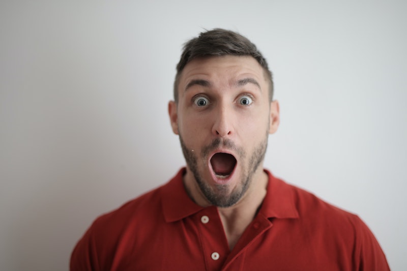 Up close image of a man with a surprised look on his face. He is wearing a red shirt and has his mouth wide open in surprise, as his eyebrows are raised high.