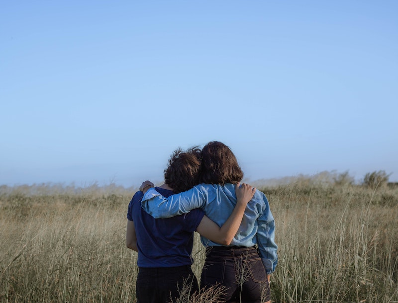 Two individuals are standing in a field under a blue sky. The individuals are facing away from the camera and each have one arm wrapped around the other. Their heads are leaning against each other.