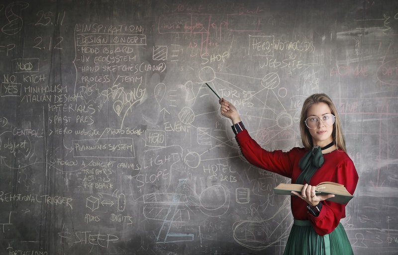 A woman wearing a red shirt and glasses is standing in front of a chalk board filled with writing and equations. The woman is holding a pencil in one hand, using it to point at the chalk board. She's holding an open book in her other hand.