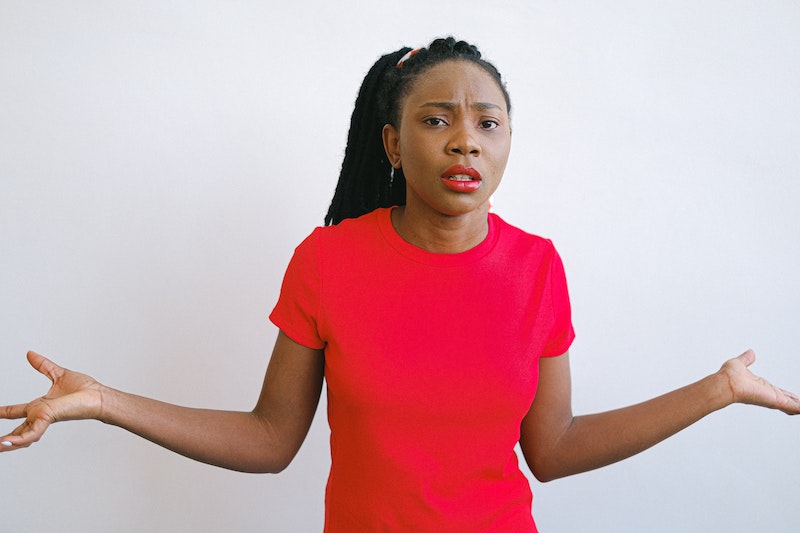 Woman is standing in front of a white wall, wearing red lipstick and a red t-shirt. She has her arms raised to her sides and a confused look on her face.