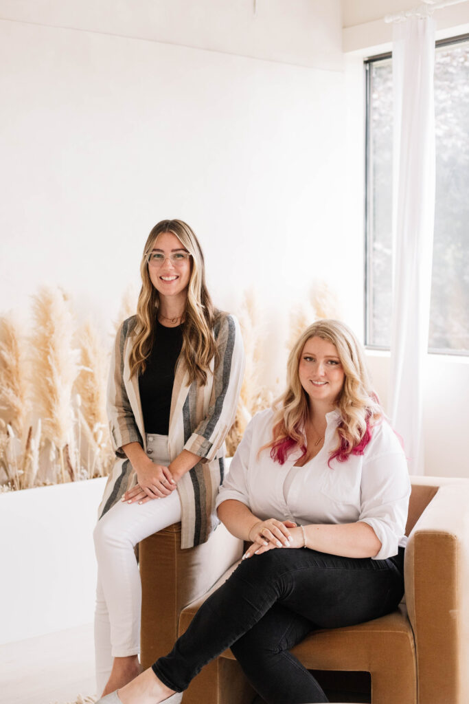 Kendra Grant and Elizabeth Adam sitting on a chair in a bright room with pampas in the background. Kendra is smiling and has long hair with glasses. Elizabeth is smiling with blonde and pink hair.