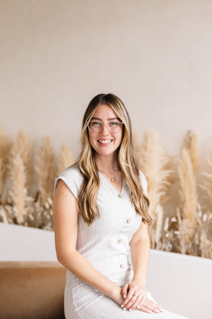 Kendra Grant is featured in this photo. She is sitting inside, with a pampas grass background. She has long hair, glasses, and is smiling in this photo.