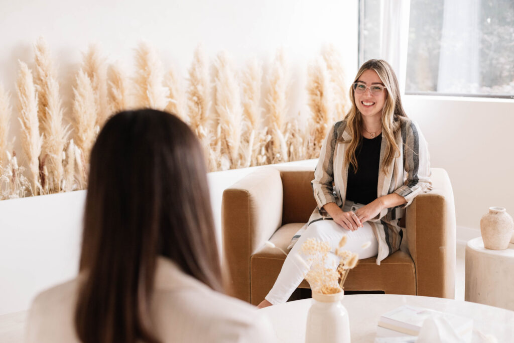 Kendra Grant sitting in a brown chair in a therapy room with a client. Kendra is smiling and listening to the client.