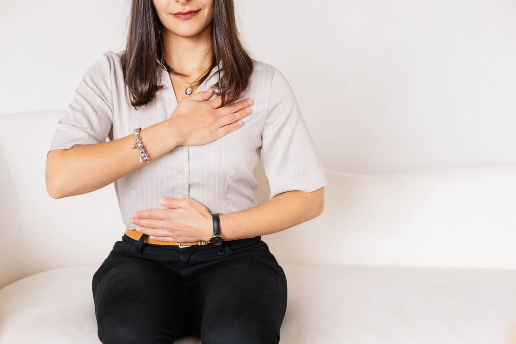Image of a women sitting in a beige room with one hand over her chest and her other arm covering her stomach. She has a soft smile. Elizabeth Adam is a registered psychotherapist providing virtual services across Ontario. She works with trauma, including a specialization in somatic experiencing.